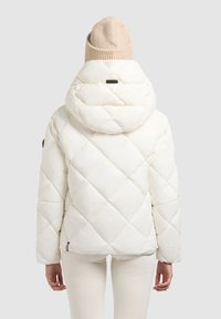 Chaqueta acolchada blanca con capucha grande, con costuras diagonales y un pequeño logo cerca del dobladillo. Llevada con pantalones en color crema.