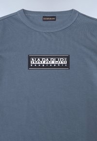Pilkos medvilnės marškinėliai su apvaliu kaklu ir stačiakampiu juodu logotipu, kuriame užrašyta "NAPAPURI geograpHIC" baltu tekstu.
