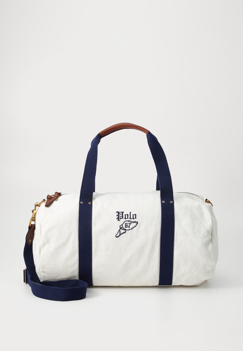 Sac de voyage en toile de couleur blanc cassé avec des anses bleu marine, garnitures en cuir, broderie du logo et une bandoulière amovible. Forme ronde avec fermeture à zip.