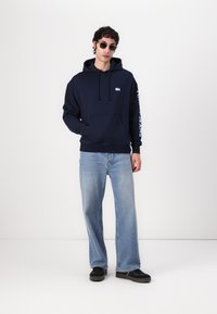 Sudadera azul marino con cordón, bolsillo frontal y logo blanco. Combinada con jeans azul claro y zapatillas negras. Lleva gafas de sol redondas.