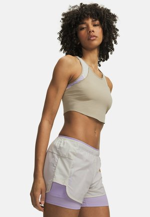 Vrouw met krullend haar, gekleed in een beige cropped tanktop en witte en lavendelkleurige gelaagde sportshorts, staand tegen een effen achtergrond.