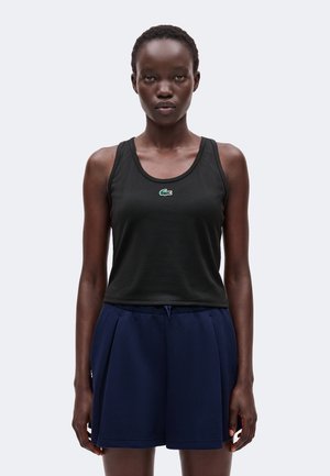 Giovane donna che indossa un top nero senza maniche con un piccolo logo verde e pantaloncini blu scuro larghi, in piedi davanti a uno sfondo semplice.