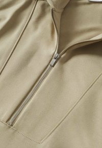 Vêtement beige avec une fermeture éclair argentée centrale partiellement ouverte, montrant des coutures détaillées et une texture de tissu lisse.