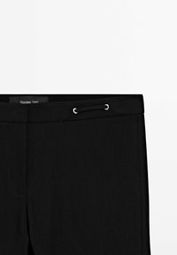 Pantalon noir sur mesure avec une fermeture frontale cachée, des œillets argentés et une petite boucle en tissu sur la ceinture.