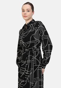 Black button-up dress featuring a geometric white line pattern, long sleeves, a waist tie, and a straight cut. Smooth fabric texture.