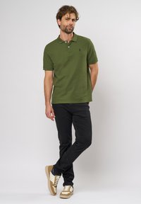Polo shirt en coton vert avec un col, arborant un petit logo sur la poitrine, associé à un pantalon noir et des baskets beige et blanches.