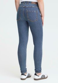 Person klädd i blå slim-fit jeans och vita sneakers med svarta r�änder, stående mot en helt ljus bakgrund.