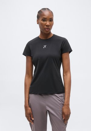 Γυναίκα με πλεξούδες στα μαλλιά, που φοράει ένα στενό μαύρο αθλητικό μπλουζάκι με κοντά μανίκια και άνετο γκρι παντελόνι, στέκεται μπροστά σε απλό ανοιχτό φόντο.