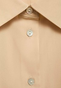 Camisa beige con cuello puntiagudo, textura suave y tres botones perlados en el frente, que exhibe sutiles detalles de costura.