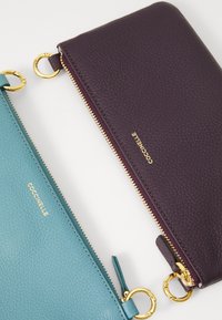 Duos tekstūruotų odinių užtrauktukų piniginių, viena turkio spalvos, kita tamsiai purpurinė, abi su auksiniais žiedais ir prekės ženklu "Coccinelle" auksinėmis raidėmis.