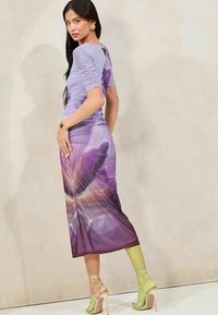 Violetti mekko, jossa on rypytetyt hihat ja kukkakuviolla varustettu liukuväri. Läpinäkyvä kangas kulkee laventelista syvään violettiin, ja se on yhdistetty vihreisiin korkokenkiin.