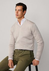 Mężczyzna siedzący na stołku, ubrany w jasnobeżowy sweter w serek na białej koszuli i oliwkowych spodniach, patrzący lekko w lewo na jednolitym tle.