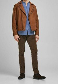Giacca biker in finta suede marrone, camicia azzurra con bottoni, pantaloni in corduroy marroni e stivali scuri. La giacca presenta cerniere e una vestibilità morbida.