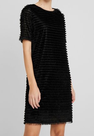 Femme portant une robe noire texturée à manches courtes avec des bandes horizontales à franges, debout devant un fond gris clair uni.