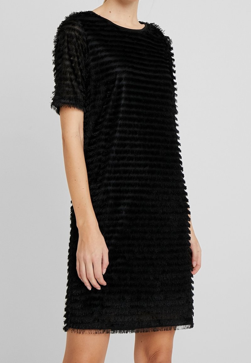 Femme portant une robe noire texturée à manches courtes avec des bandes horizontales à franges, debout devant un fond gris clair uni.