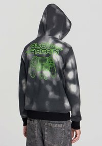 Hoodie in tie-dye bianco e nero con un controller di gioco in wireframe verde neon e la scritta "Play Again" sul retro, abbinato a jeans grigi.