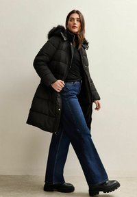 Długa czarna puchowa kurtka z kapturem podbitym futrem, w zestawieniu z czarną bluzką i szerokimi niebieskimi dżinsami. Noszona z czarnymi botkami na grubej podeszwie.