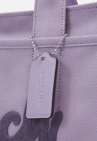 Violetti kangaskassi teksturoidulla pinnalla, kirjailtu logomalli ja hopeinen helmiketjulla kiinnitetty roikkuva tunniste.