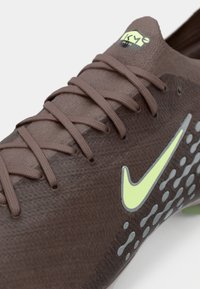 Καφέ αθλητικό παπούτσι με συνθετική υφή, διαθέτει γκρι κουκίδες και νεον κίτρινο λογότυπο Nike στο πλάι. Περιλαμβάνει επίπεδα κορδόνια.