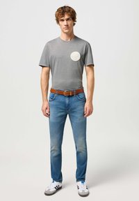 T-shirt grise à manches courtes avec un col rond et un écusson logo circulaire, associée à un jean bleu, une ceinture marron et des chaussures blanches avec des rayures noires.