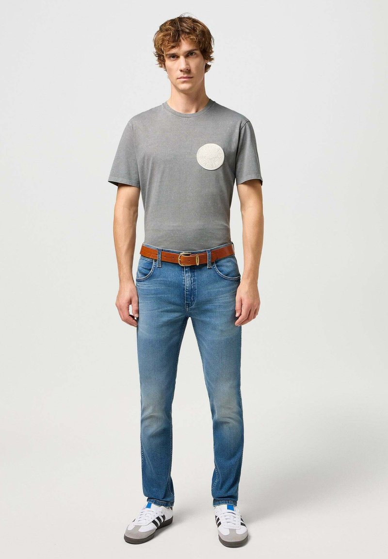 T-shirt grise à manches courtes avec un col rond et un écusson logo circulaire, associée à un jean bleu, une ceinture marron et des chaussures blanches avec des rayures noires.