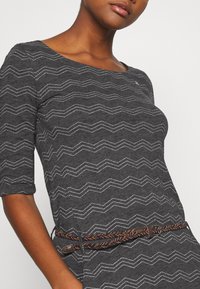Graues Kleid mit einem Zickzackmuster und weißen Akzenten, 3/4-Ärmeln und einem gewebten braunen Gürtel, der die Taille betont.