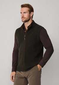 Zelená pletená vesta s vysokým límcem, zipovým zapínáním a žebrovanou strukturou, kombinována s tmavě hnědým tričkem s dlouhým rukávem a béžovými kalhotami.