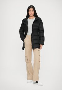 Svart boblejakke med hette, med glidelås og horisontalt quiltet design. Kombinert med beige cargo-bukser og hvite joggesko.