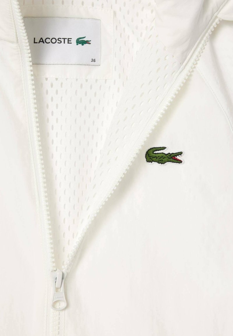 Biała kurtka na zamek błyskawiczny z siateczkową podszewką, z zieloną haftowaną aplikacją w kształcie krokodyla oraz teksturowanym materiałem. Zamek błyskawiczny z uchwytem oznaczonym "LACOSTE."