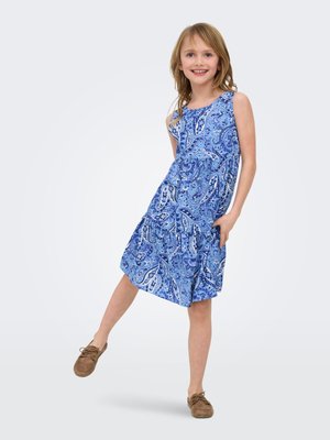 Smiling girl in sleeveless blue paisley dress poses with one leg raised, wearing brown lace-up shoes against a white background.