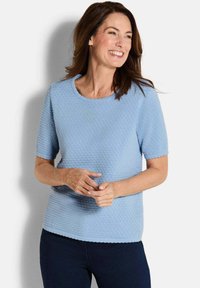 Vrouw die glimlacht, gekleed in een textuur lichte blauwe korte mouwen top en donkere broek, staande tegen een effen witte achtergrond.