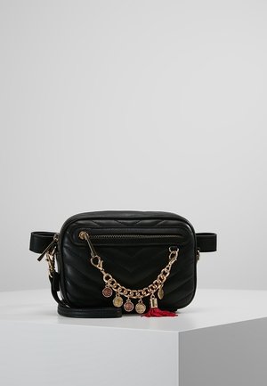 Zwart gewatteerde crossbodytas met voorvak met rits, gouden ketting met muntbedels en rood kwastdetail, tentoongesteld op een witte ondergrond.