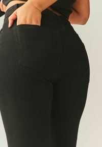 Juodos spalvos „bootcut“ džinsai su lygia tekstūra, turintys dvi galines kišenes, vidutinio aukščio liemenį ir minimalistinius siuvimo detalių elementus.