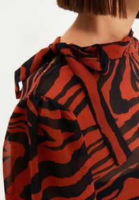 Punainen ja musta tiikeriraitainen pusero, jossa solmittava kaulus. Läpinäkyvä kangas, liehukkeet hihat ja teksturoitu viimeistely korostavat sen muotoilua.