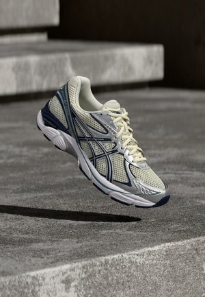 Beige og navy løbesko med mesh og syntetisk overdel, snørebånd er bundet, hænger over betonstrappen.