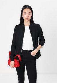 Veste bomber noire avec fermeture éclair, poignets à côtes, portée par-dessus un haut blanc à côtes. Le modèle tient un petit sac à main rouge. Un jean noir complète le look.