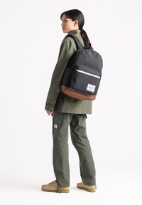 Zaino nero e marrone con una tasca frontale e zip bianca, accanto a una persona in un outfit verde oliva e scarpe beige.