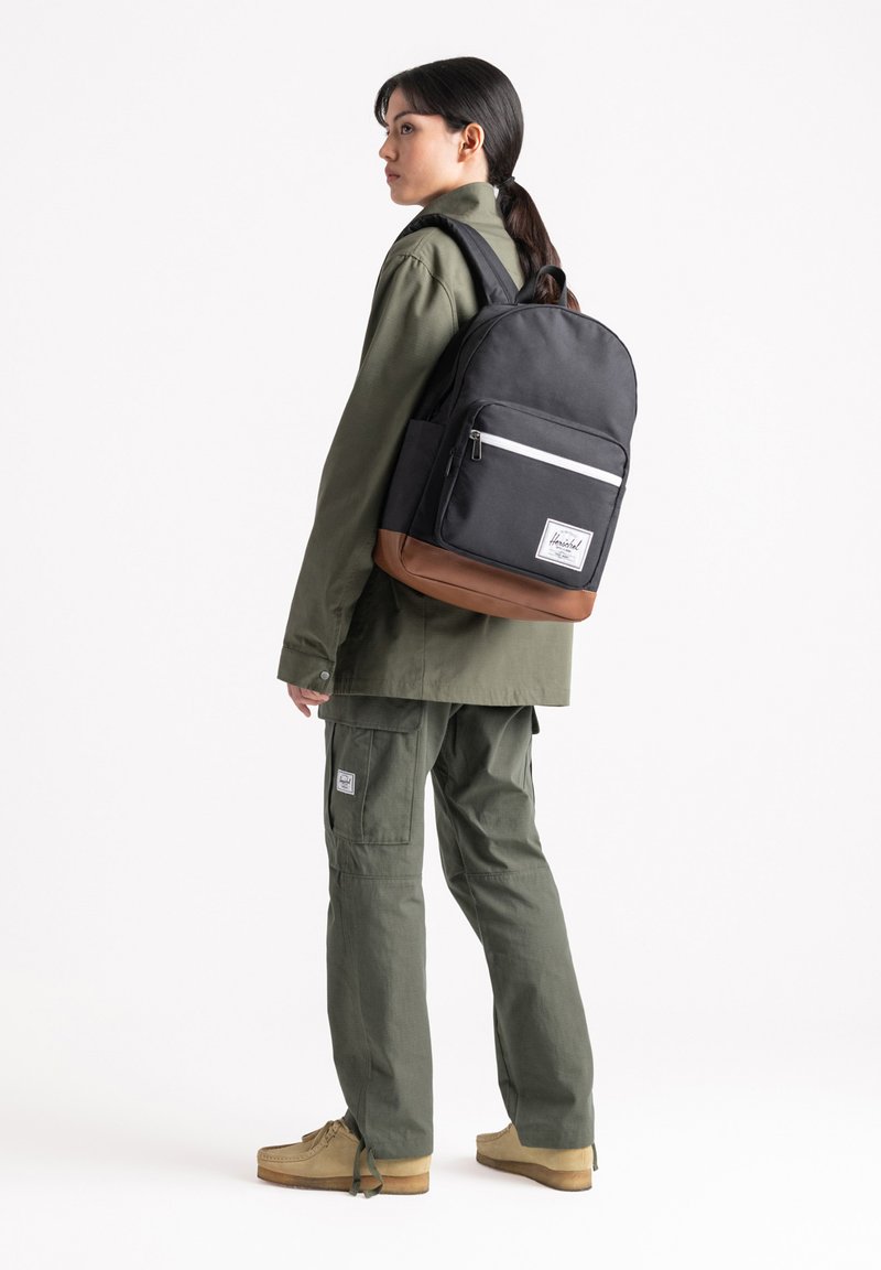 Zaino nero e marrone con una tasca frontale e zip bianca, accanto a una persona in un outfit verde oliva e scarpe beige.