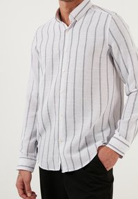Bílá košile s dlouhými rukávy a námořnickými vertikálními pruhy, vyrobená z texturovaného materiálu, s límečkem na knoflíky a diskrétními manžetami.