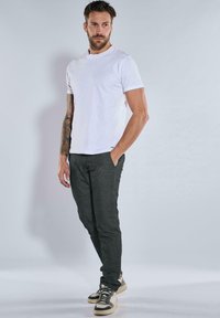 White crew-neck t-shirt, dark gray fitted pants, and patterned sneakers. Model has tattoos and stands with one hand in the pocket.