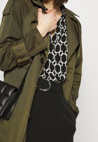 Oliwkowy trencz z paskiem w talii, nałożony na bluzkę w czarno-białym wzorze łańcucha, zestawiony z czarnymi spodniami o wysokim stanie i czarną torebką.