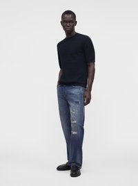 Homme debout portant un pull noir à manches courtes, un jean bleu déchiré et des chaussures noires sur un fond gris clair uni.