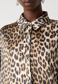 Femme portant une chemise à imprimé léopard avec des boutons à motif floral et des boucles d'oreilles créoles argentées, montrée du cou à la poitrine.