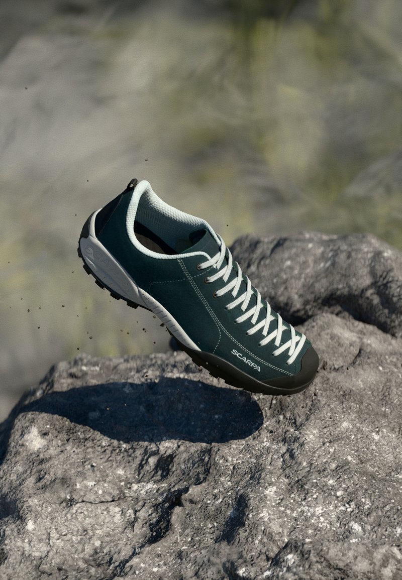 Shoe de camurça verde-escura com atacadores brancos, biqueira de borracha e sola texturizada, suspenso acima de uma superfície de rocha áspera.