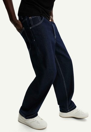Dark blue denim jeans with a relaxed fit, featuring white stitching, side pockets, and a contrasting waistband. Paired with white sneakers.