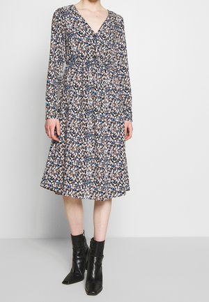 Femme portant une robe fleurie à manches longues, col en V, longueur au genou, avec des motifs noirs, blancs et bleus, associée à des bottines noires.