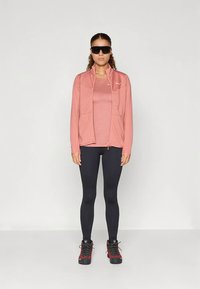 Žena u ružičastoj jakni i majici, crnim tajicama, crvenim planinarskim čizmama i crnim sunčanim naočalama, stoji na jedinstvenoj bijeloj pozadini.