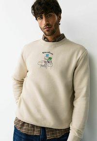 Beiger Sweatshirt mit gerippten Bündchen, auf der Vorderseite mit einer Grafik der Cartoonfiguren "Wallace & Gromit". Darüber getragen ein kariertes Hemd.
