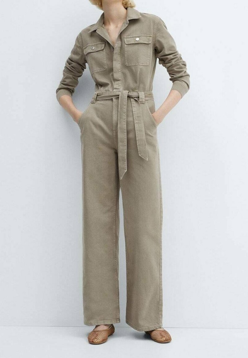 Combinaison en khaki à manches longues, avec des poches poitrine boutonnées, une taille ceinturée, et un pantalon large. Tissu lisse et design minimaliste.