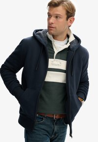 Marineblå hættetrøje med snore, grå inderfor og sidelommer, lagdelt over en grøn polo med hvide striber. Brun bæltet og denim jeans.